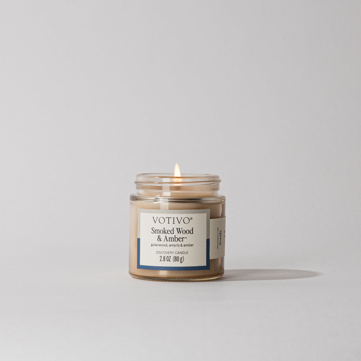 Votivo Smoked Wood & Amber Aromatic Candle, 2.8 oz, in a clear glass jar with a lit wick on a white background.