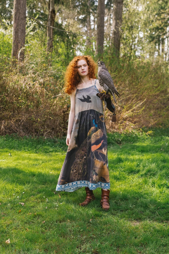 Woman in Market of Stars Wild Beauty Bohéme Slip Dress with bird & peacock print, holding a falcon in a forest.