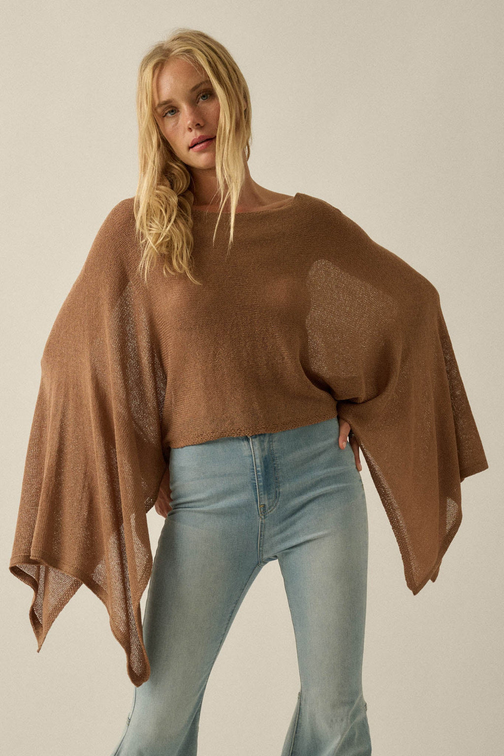 Blonde woman in Promesa Sheer to Stay Knit Poncho Sweater in Taupe with wide sleeves and light blue jeans.