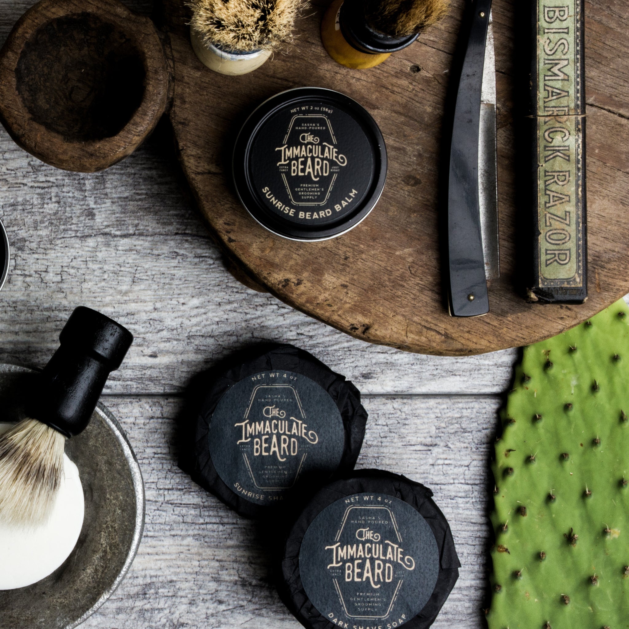 The Immaculate Beard Dark Shave Soap puck with balm, straight razor, and shaving brush on wood.