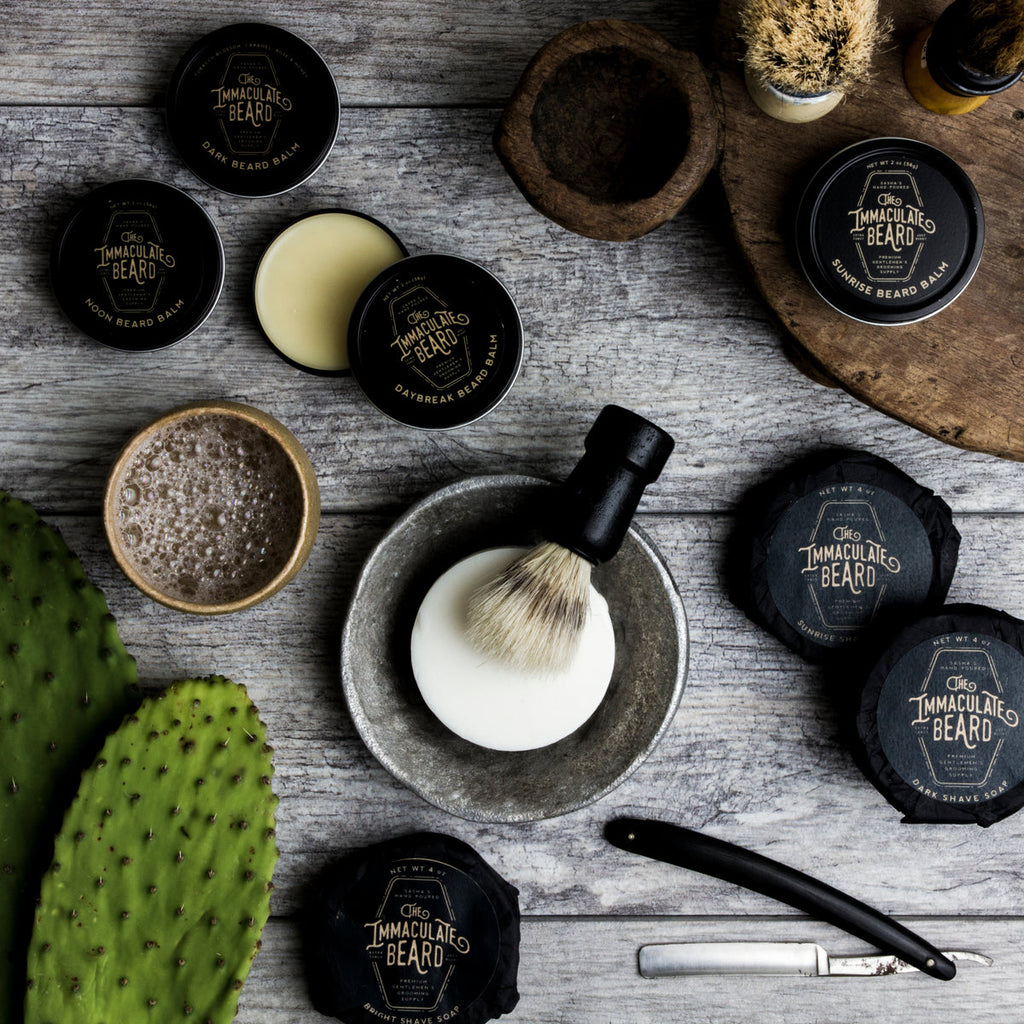 The Immaculate Beard Midnight Shave Soap Puck, with shaving brush, straight razor, and beard balms on a wooden table.