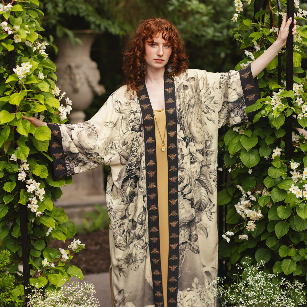 Honey & The Moon Opera Duster by Market of Stars. Woman in a cream duster with black floral and bee prints in a garden.