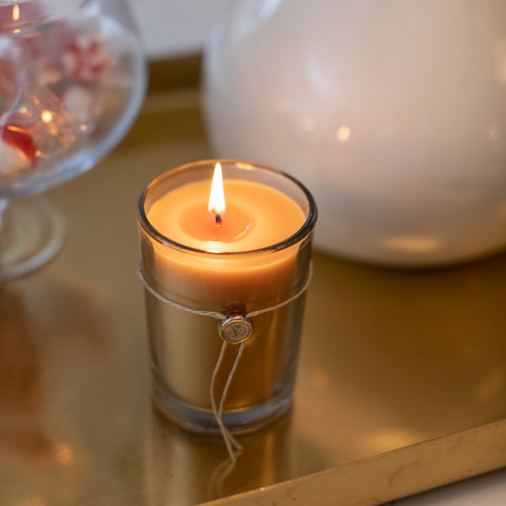 Votivo Champaca Aromatic Candle, lit, in a glass holder with twine and a seal, on a golden tray. Warm glow.
