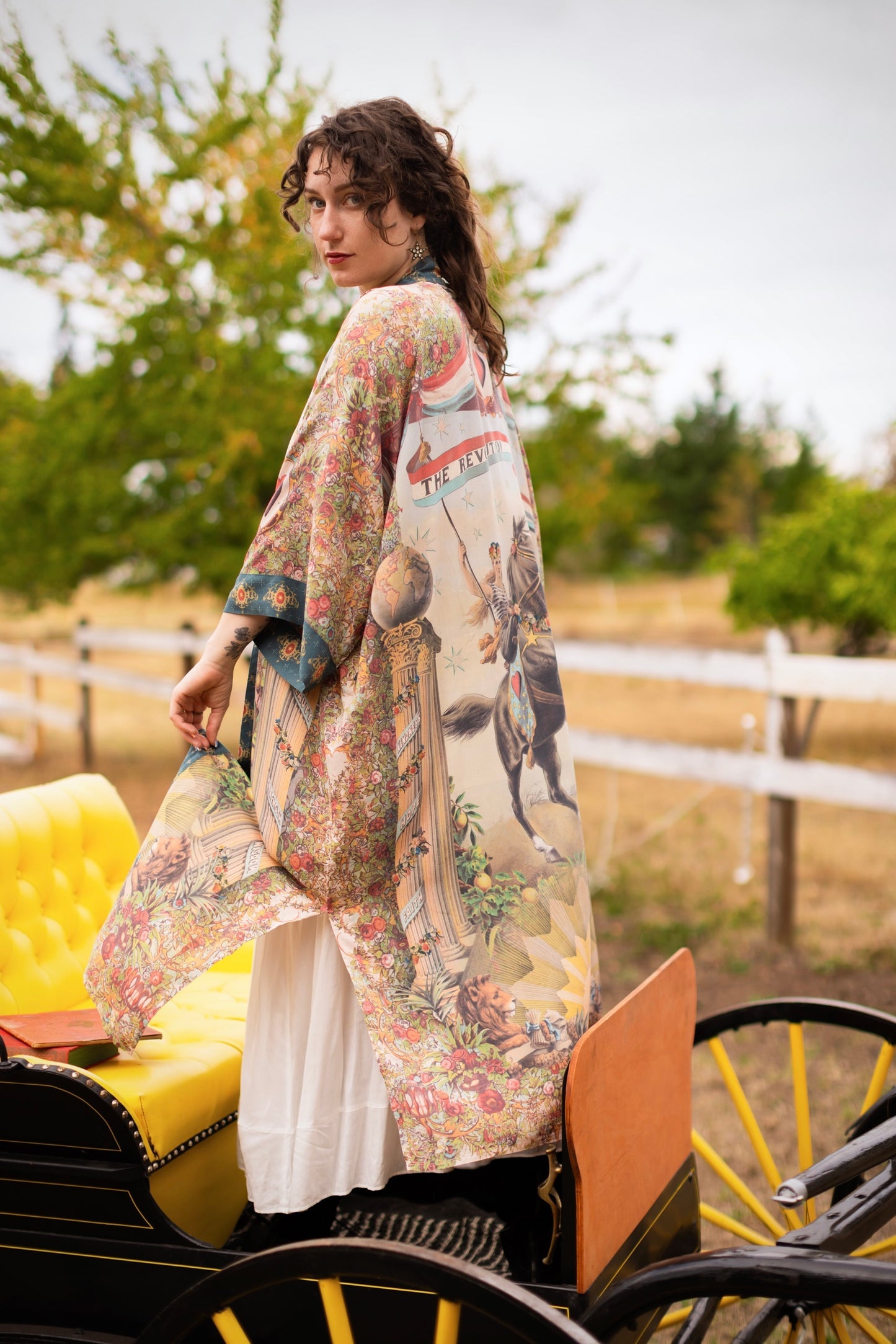 Woman in Market of Stars Love Revolution Opera Duster with vibrant, intricate prints featuring horses, lions, and florals, standing by a vintage carriage.