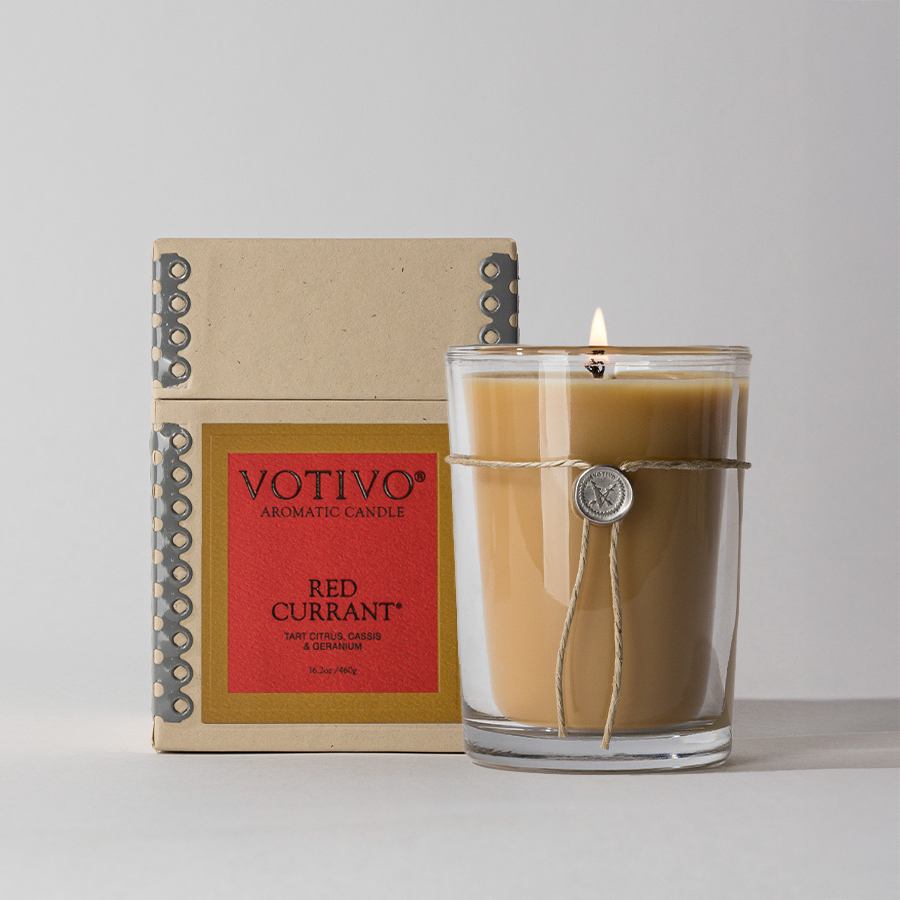 Votivo Red Currant Aromatic Candle, lit, in glass jar next to its open box. Tart citrus, cassis & geranium scent.