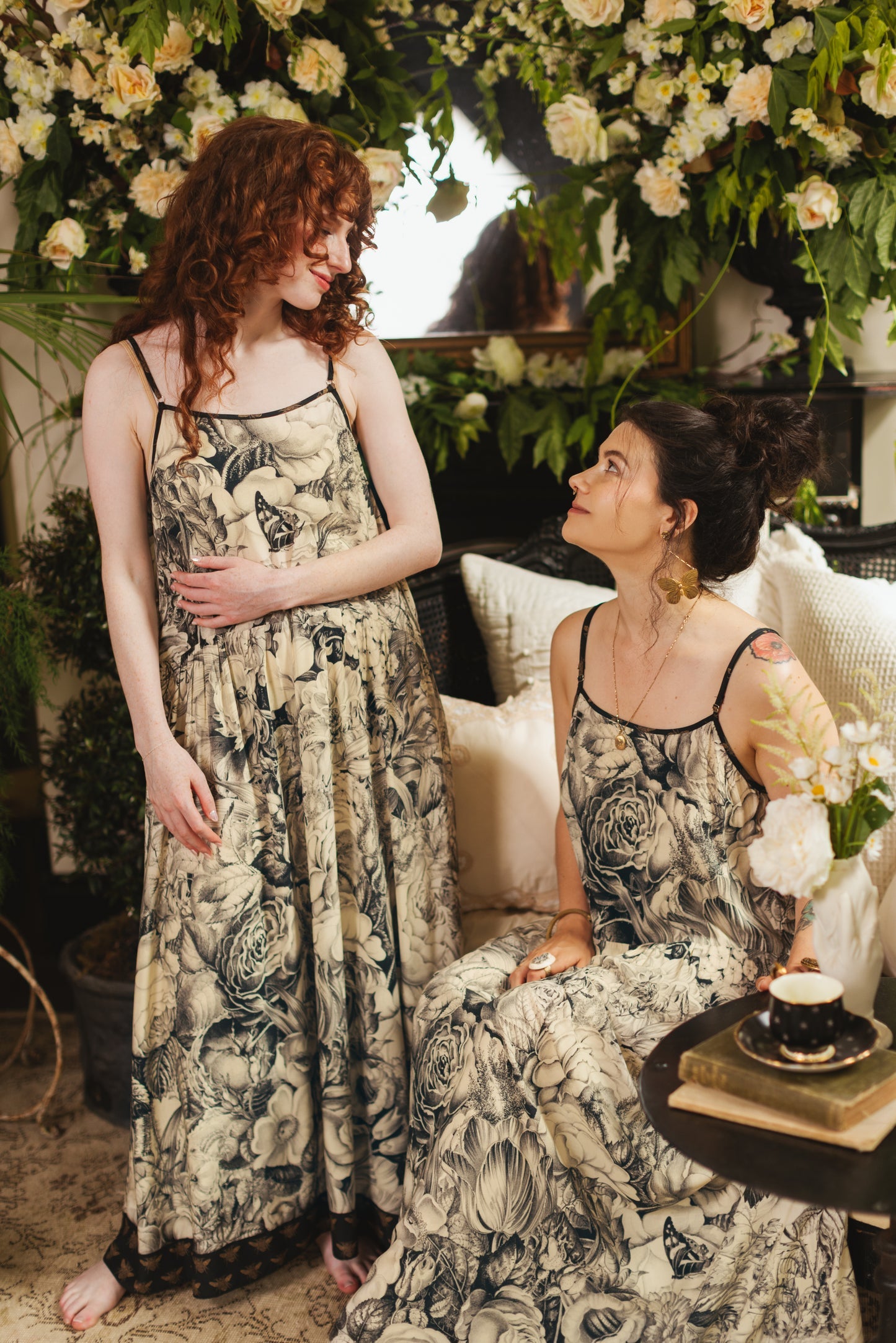 Honey & The Moon Bohéme Slip Dress | Market of Stars. Two women in matching elegant floral slip dresses surrounded by lush flowers.
