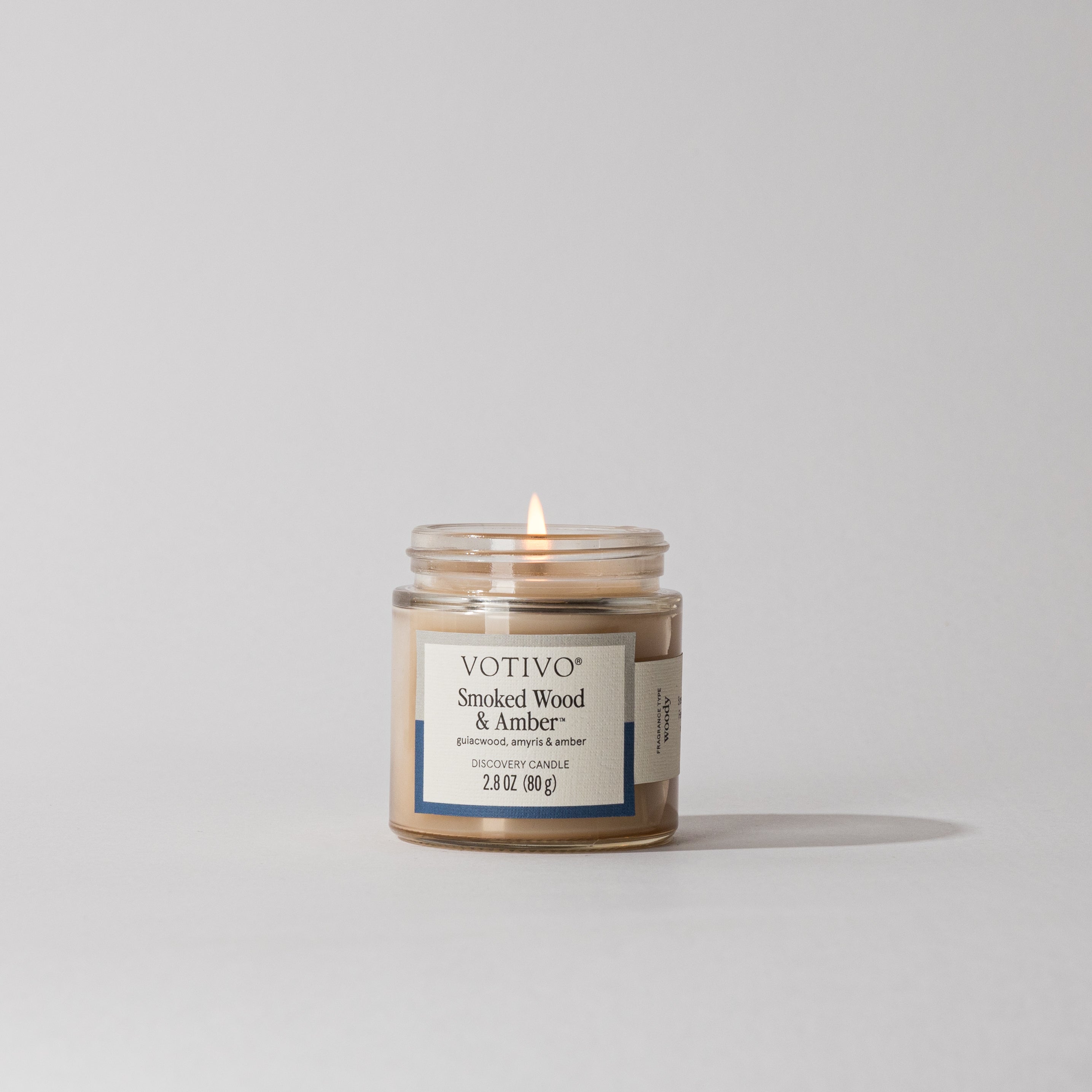 Votivo Smoked Wood & Amber Discovery Candle lit, in a clear glass jar, on a white background. Guaiacwood, amyris, amber.