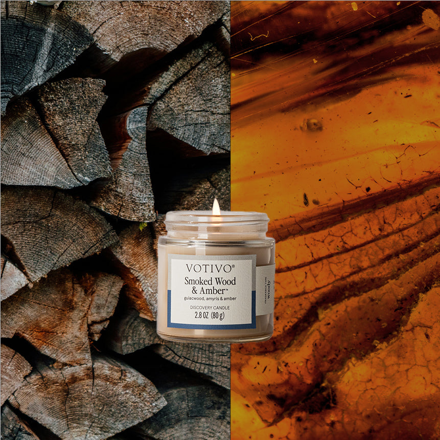 Votivo Smoked Wood & Amber Discovery Candle, burning, with a split background of stacked firewood and amber.