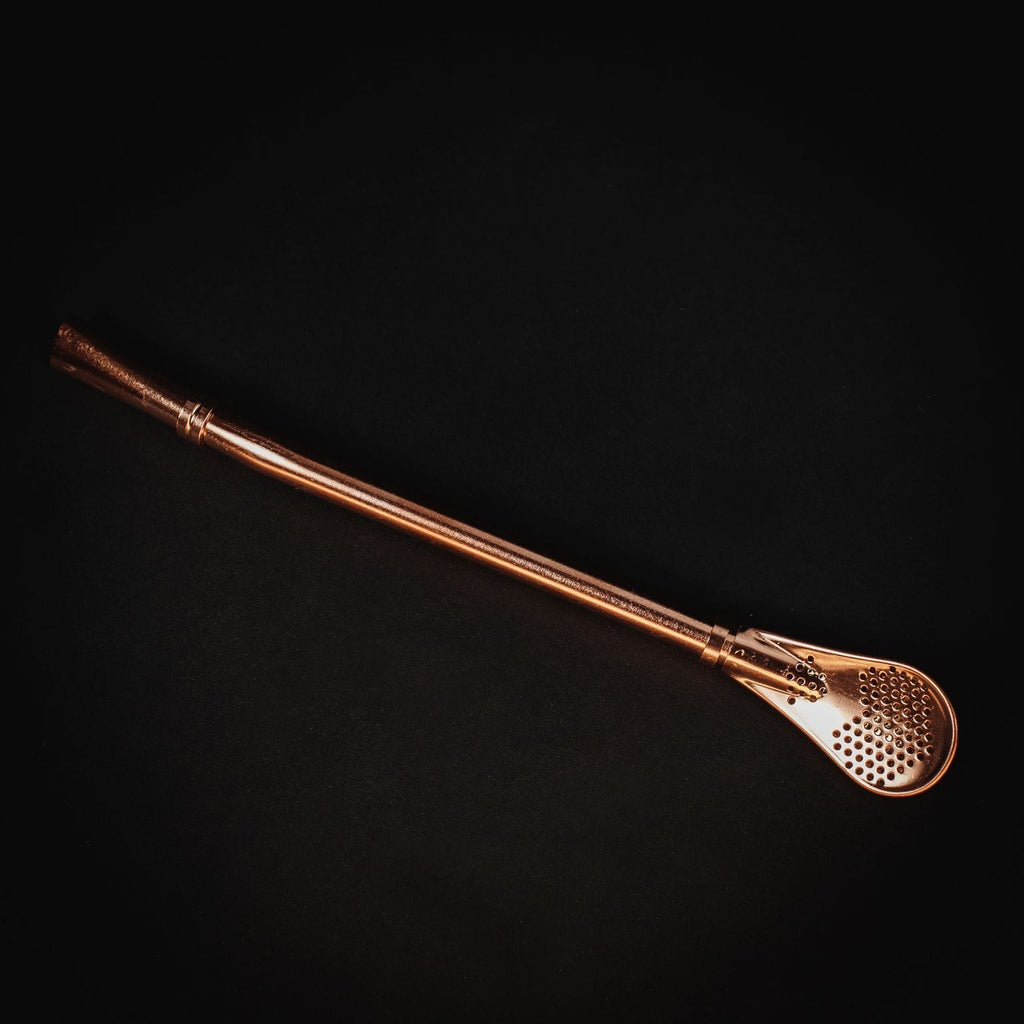 Gold Sips Tea Straw & Strainer Bombilla by Magic Hour, a golden metal straw with a filter, on a black surface.