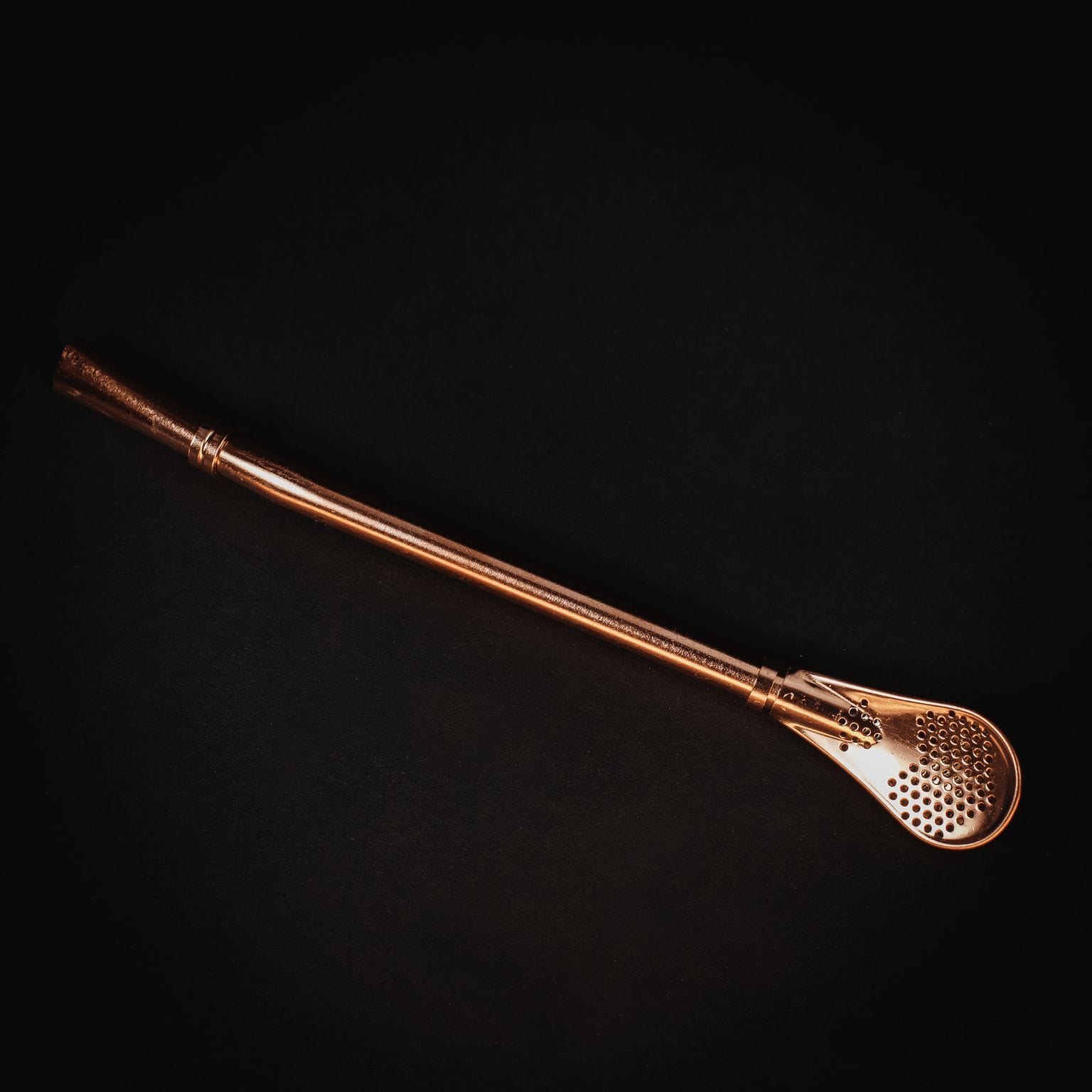 Gold Sips Tea Straw & Strainer Bombilla by Magic Hour, a golden metal straw with a filter, on a black surface.