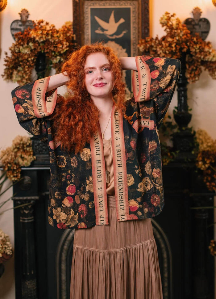 Woman in Heart of Gold Pixie Duster from Market of Stars, a dark floral kimono with 'LOVE & TRUTH FRIENDSHIP' trim, smiling.