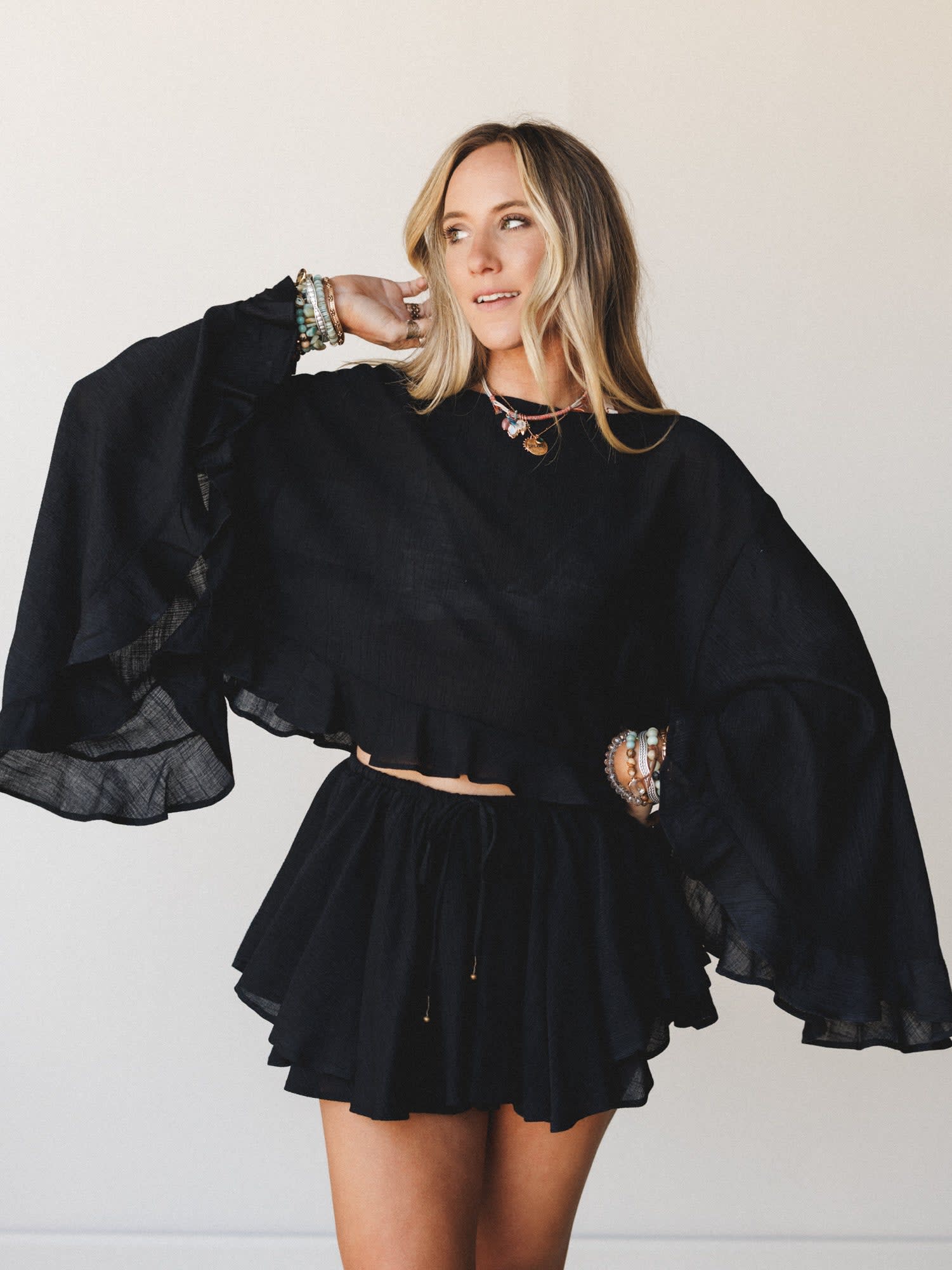 L9403T Cropped Blouse in Black by By Together, modeled with matching ruffled mini skirt and jewelry.
