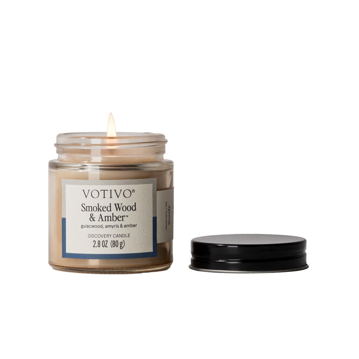 Lit Votivo Smoked Wood & Amber Discovery Candle (2.8 oz) in a glass jar with its black lid on a white background.