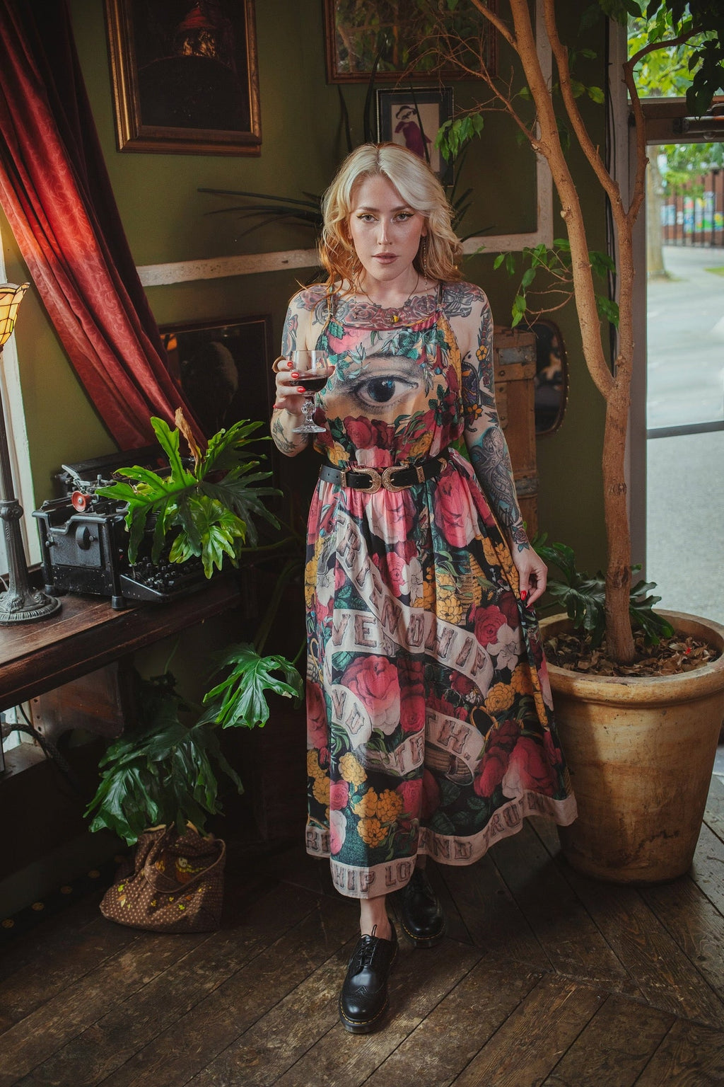 Woman in 'Friendship Love & Truth Bohéme Slip Dress' with eye & floral print, holding wine in a vintage room with plants.