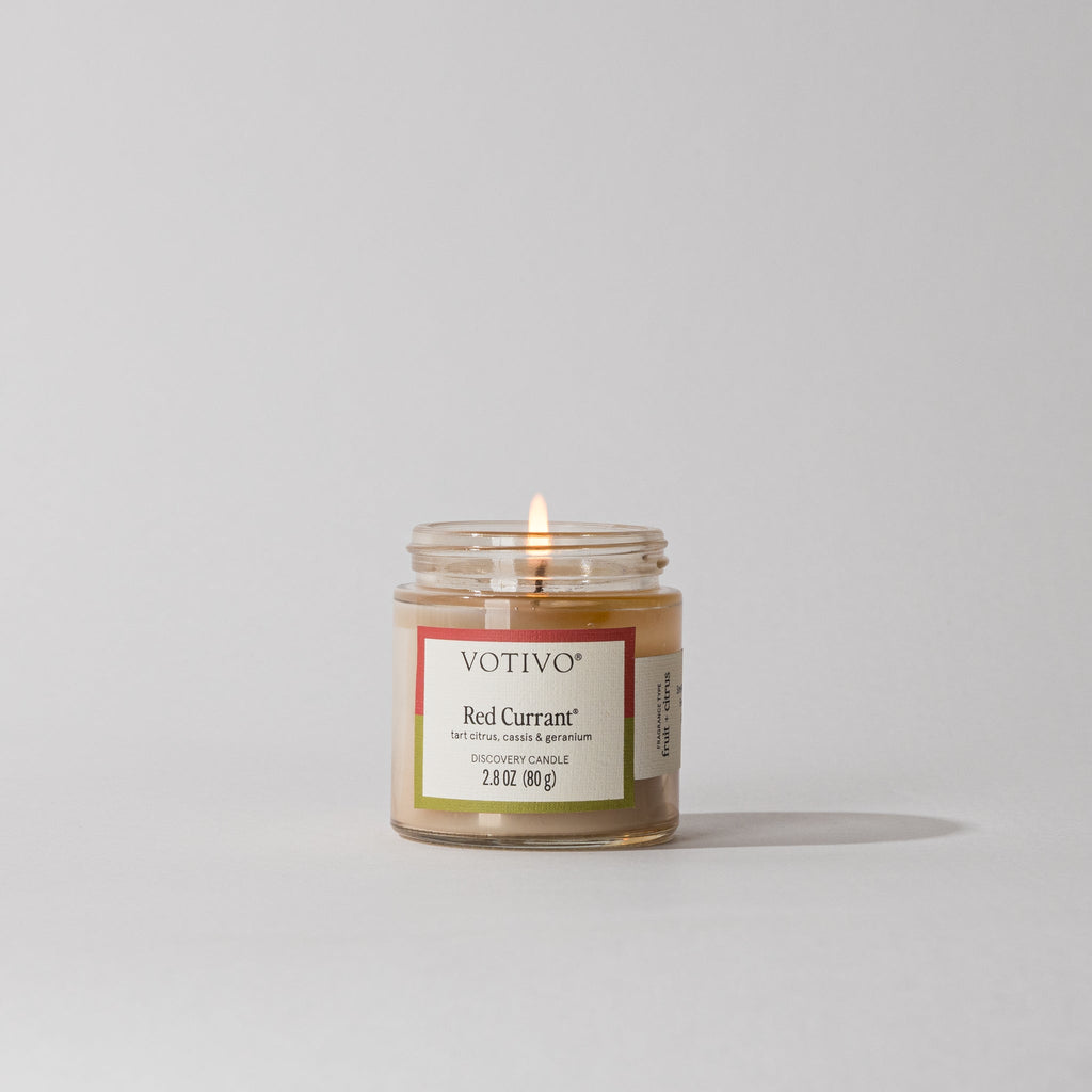 Votivo Red Currant Discovery Candle, 2.8 oz, tart citrus, cassis & geranium, burning wick, in a clear glass jar on white background.