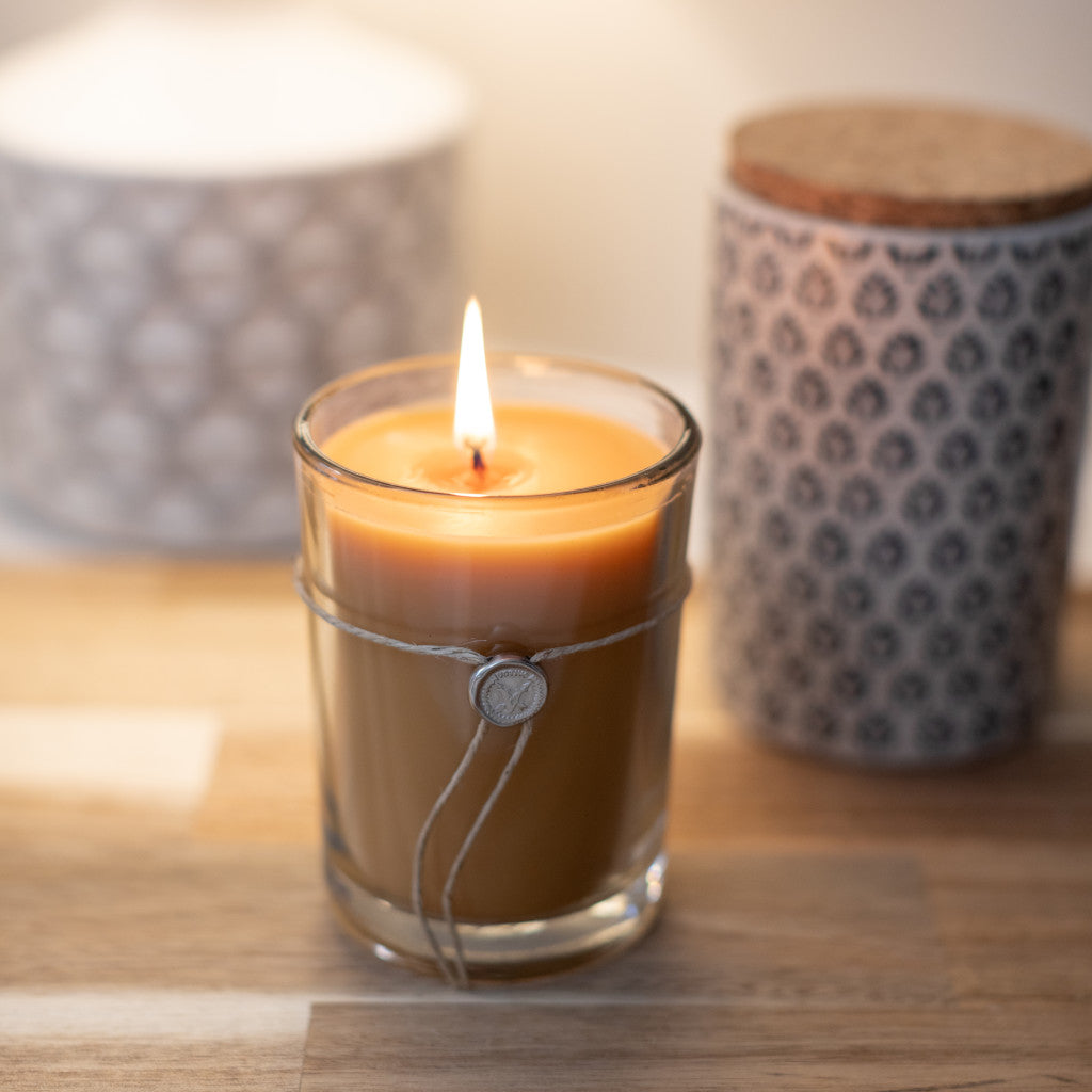Votivo Champaca Aromatic Candle, burning in a clear glass jar with twine detail, on wood, with blurred decorative jars.