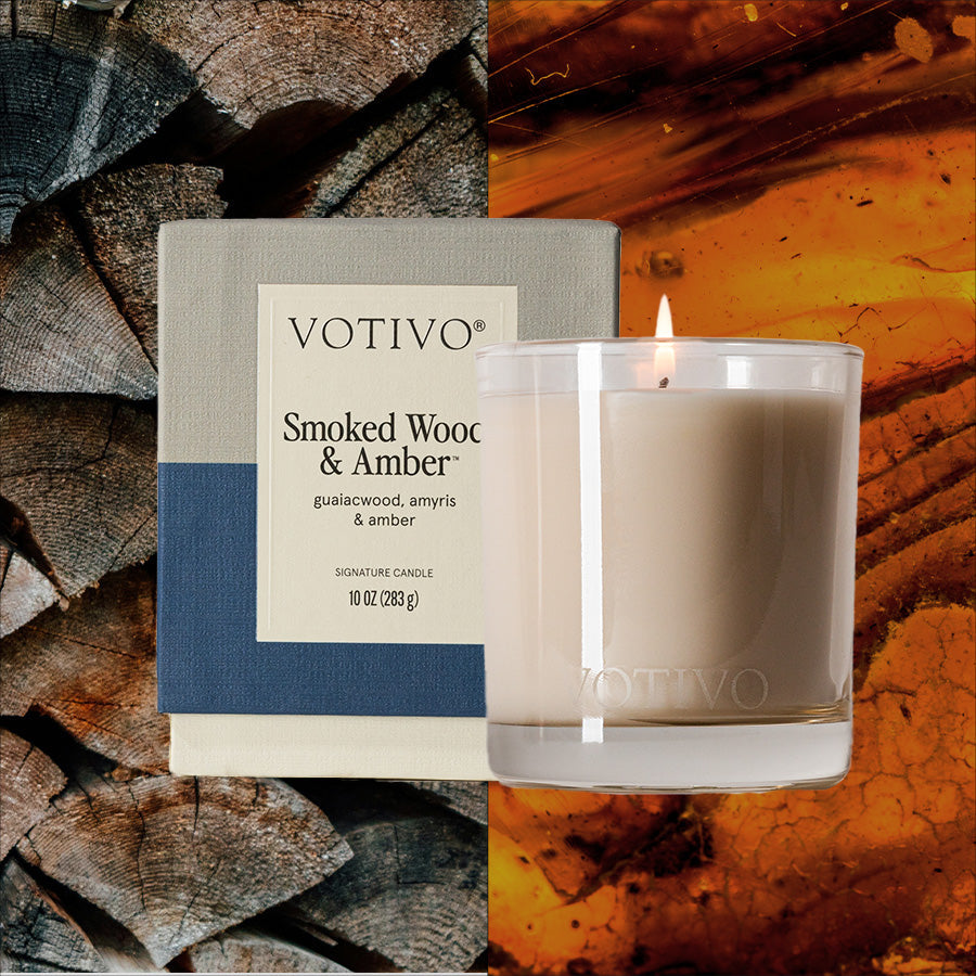 Votivo Smoked Wood & Amber aromatic candle in a glass holder, lit, with its packaging, against rustic wood and amber.