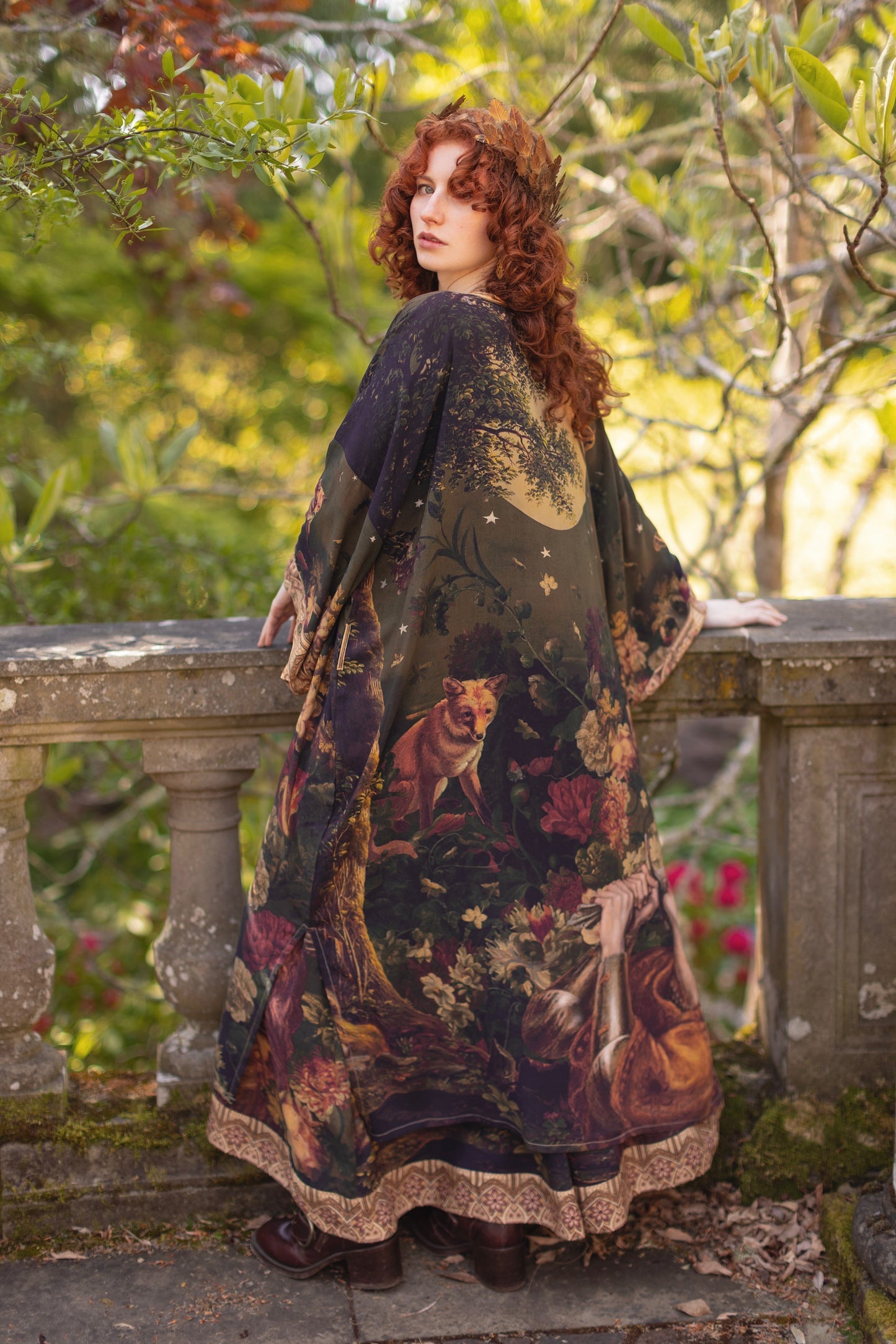 Woman with red hair and leaf crown wearing Wild Beauty Opera Duster by Market of Stars, featuring fox, deer, and floral print, standing on a stone balcony.