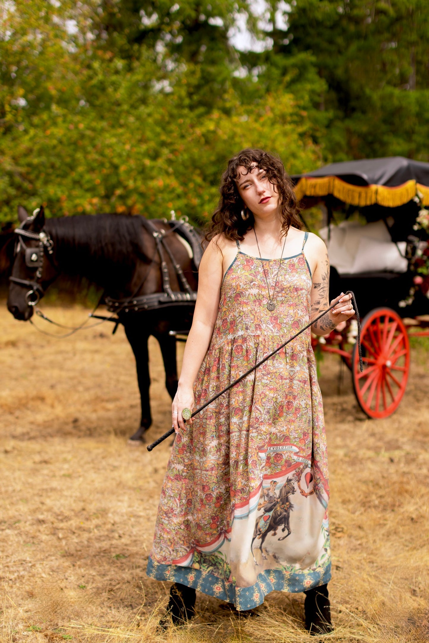 Woman in Market of Stars Love Revolution Bohéme Slip Dress, holding a riding crop, with a horse and carriage behind her.