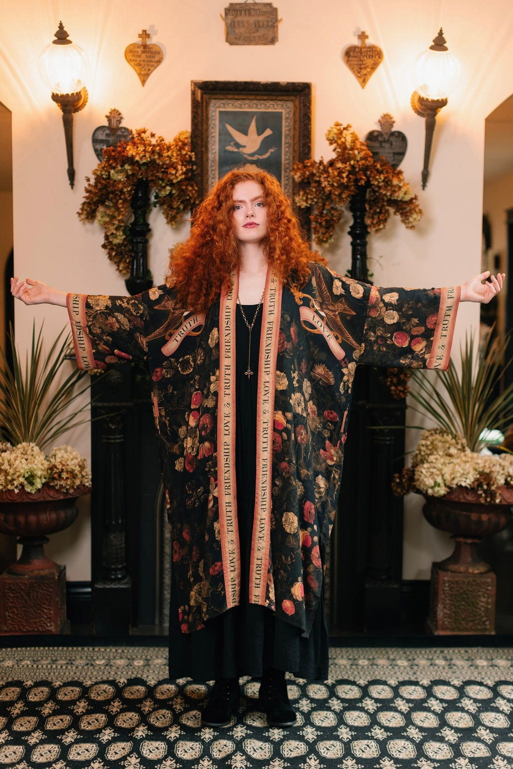 Red-haired woman in Market of Stars Heart of Gold Opera Duster with floral print and 'TRUTH', 'LOVE' text, arms outstretched.