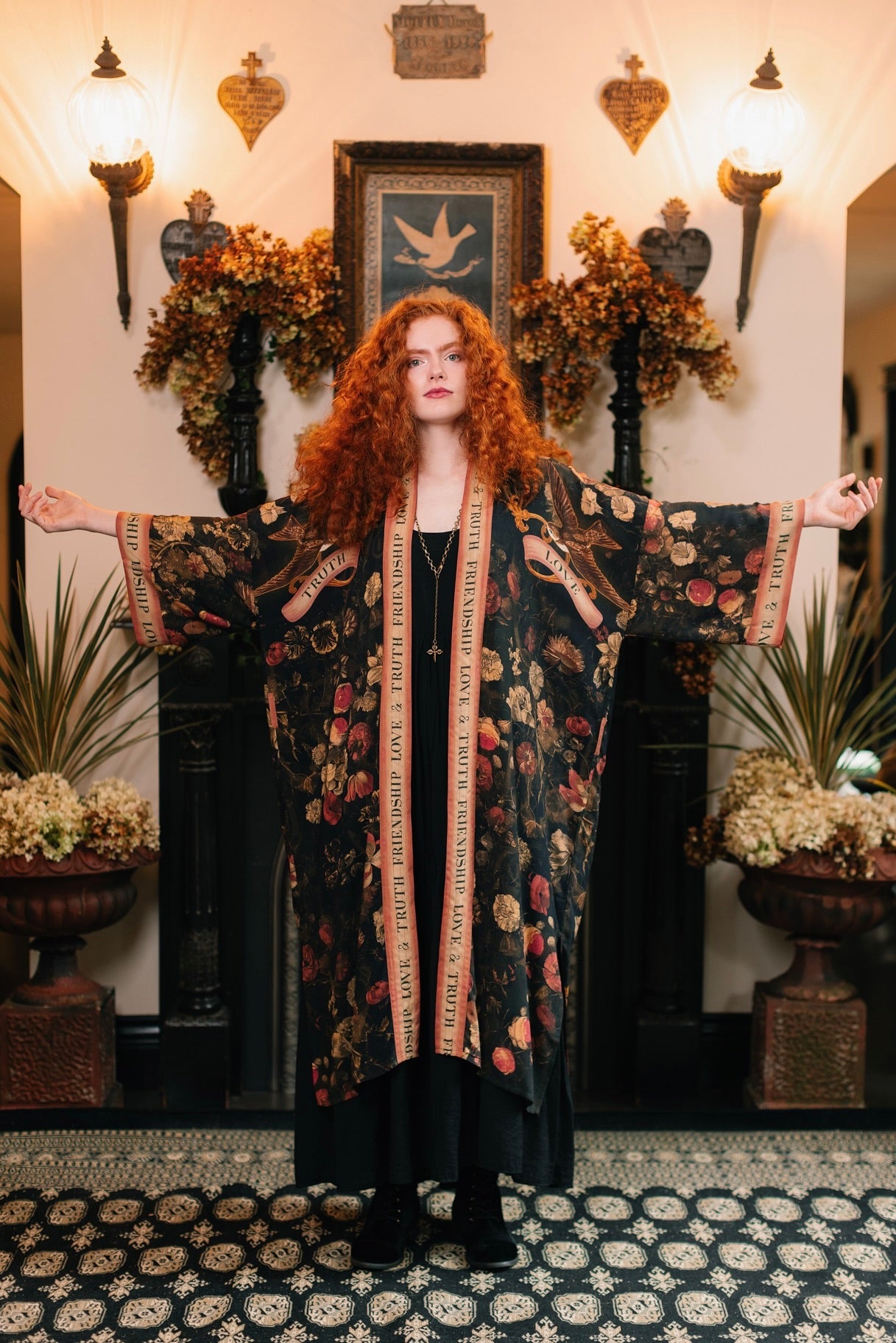 Red-haired woman in Market of Stars Heart of Gold Opera Duster with floral print and 'TRUTH', 'LOVE' text, arms outstretched.
