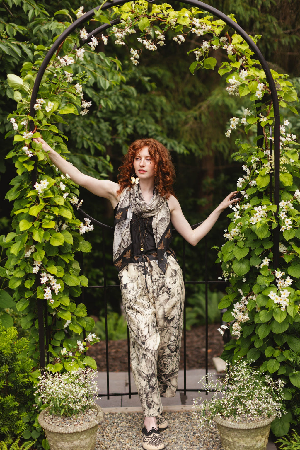 Woman in Honey & The Moon Artist Pants from Market of Stars & matching scarf, standing under a white floral archway in a garden.