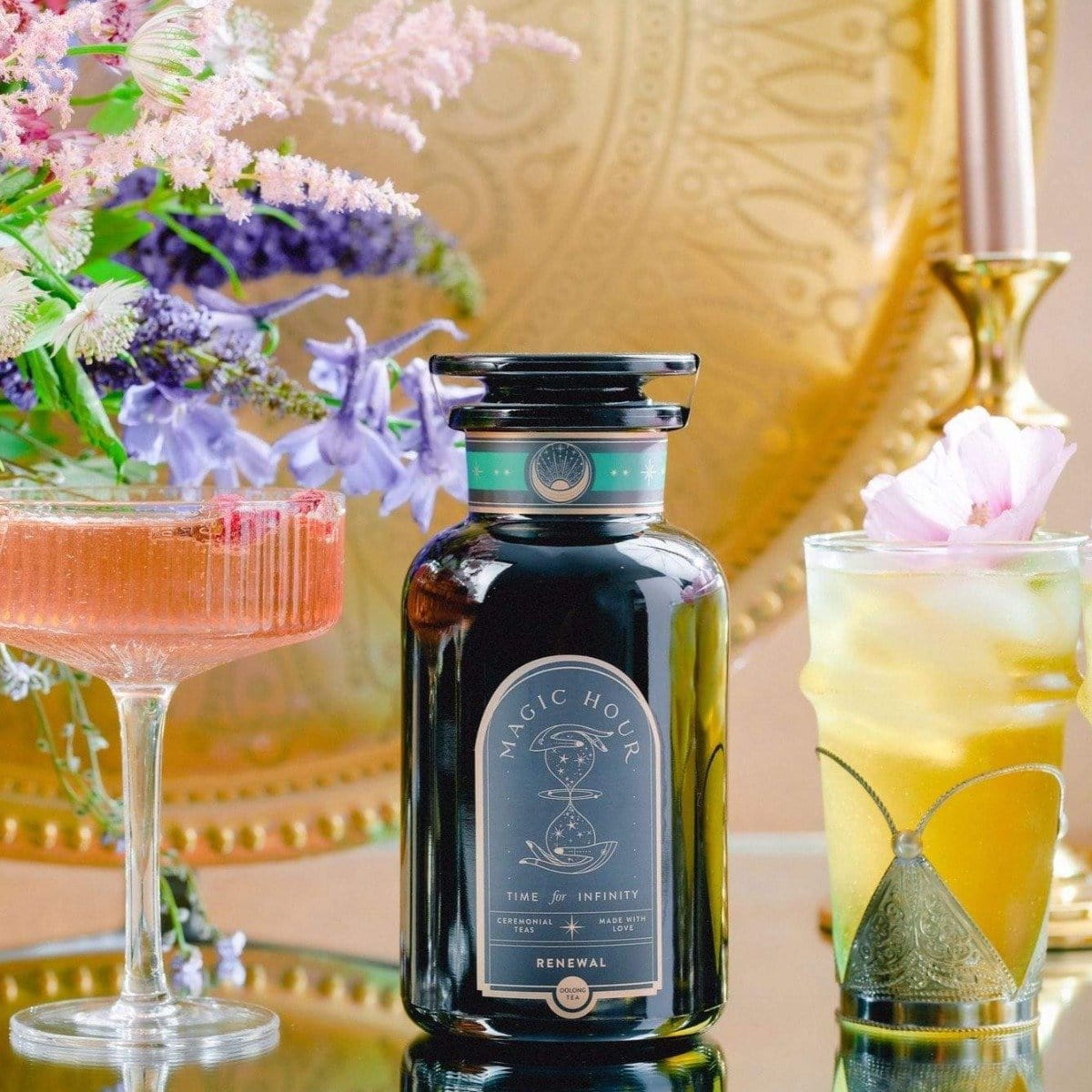 Magic Hour Renewal: Peach-Goji-Rose Oolong Tea bottle amidst two cocktails and floral arrangements on a golden tray.