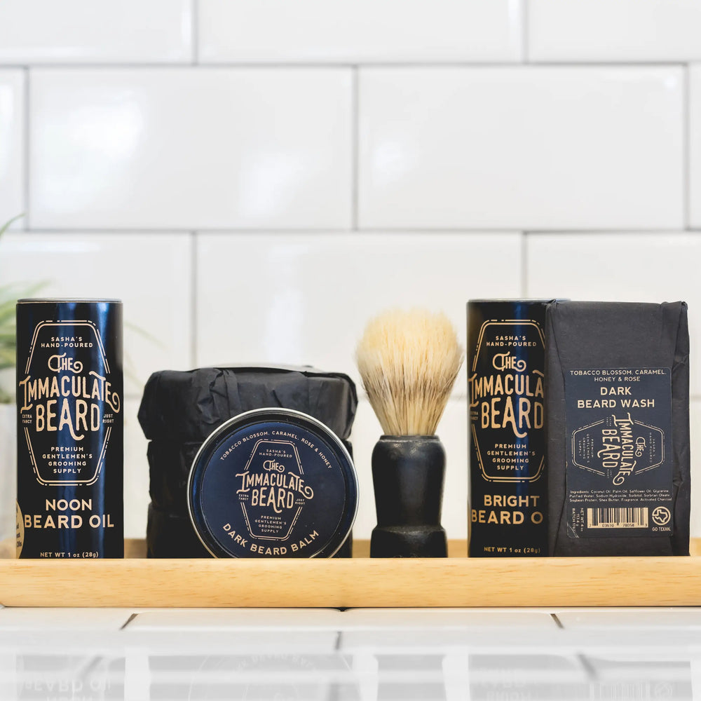 The Immaculate Beard Dark Shave Soap Puck, beard oil, balm, wash, & brush on a wooden tray against white tiles.