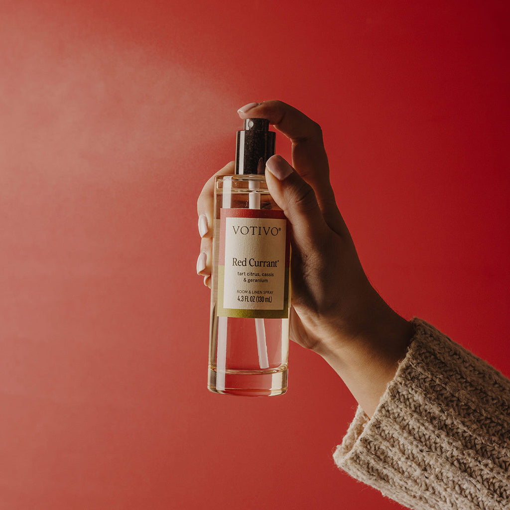 Hand holding and spraying Votivo Red Currant Room & Linen Spray bottle against a vibrant red background.