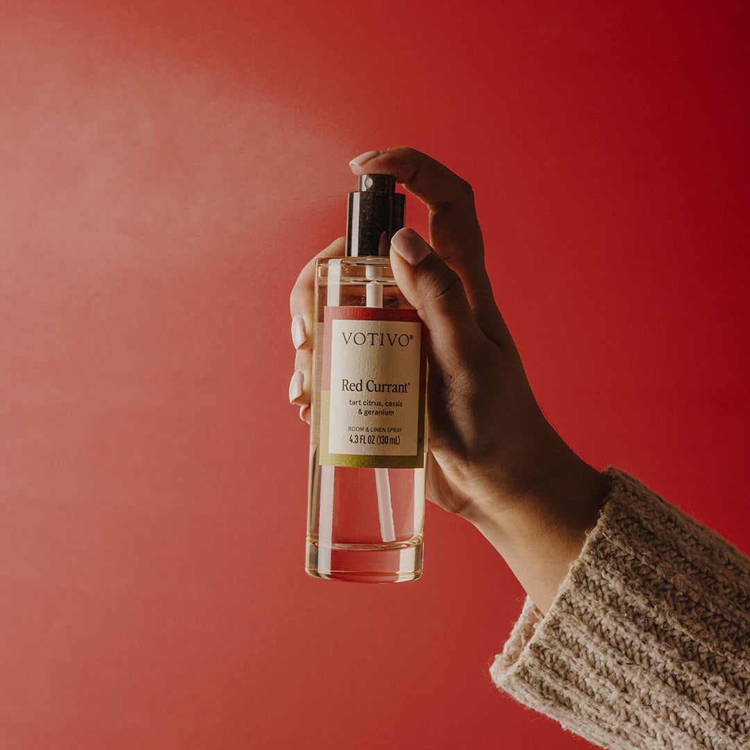 Hand holding and spraying Votivo Red Currant Room & Linen Spray bottle against a vibrant red background.