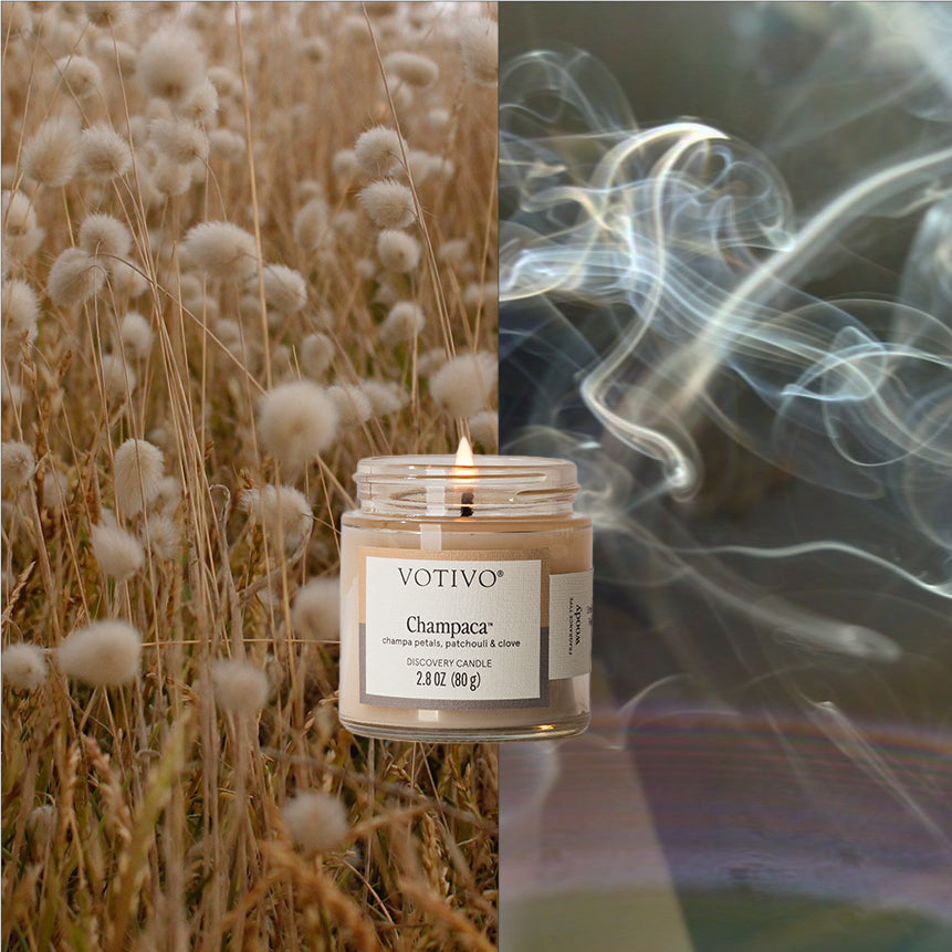 Votivo Champaca Discovery Candle burning, with a background of fluffy brown plants and swirling white smoke.