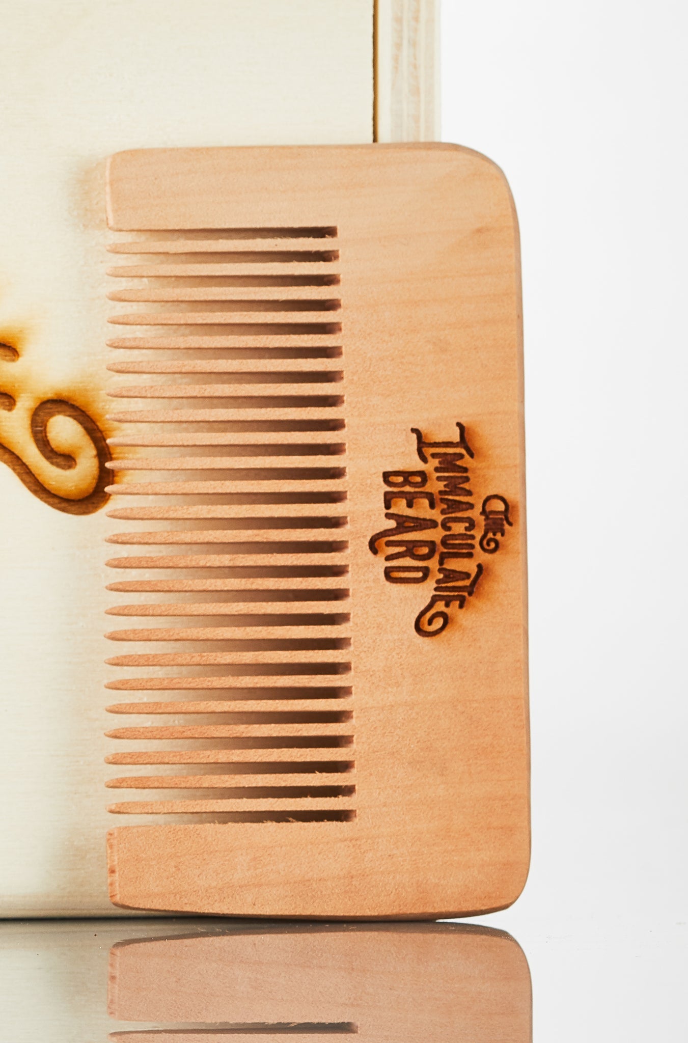 Wooden Beard Comb from The Immaculate Beard, featuring its engraved logo, resting on a reflective surface.