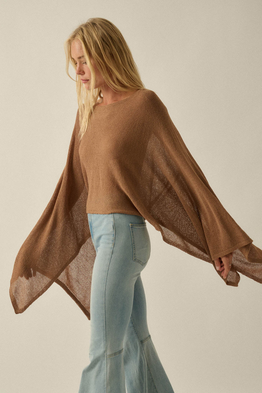 Blonde woman wearing a Promesa Sheer to Stay Knit Poncho Sweater in Taupe with flowing sleeves and light wash jeans.