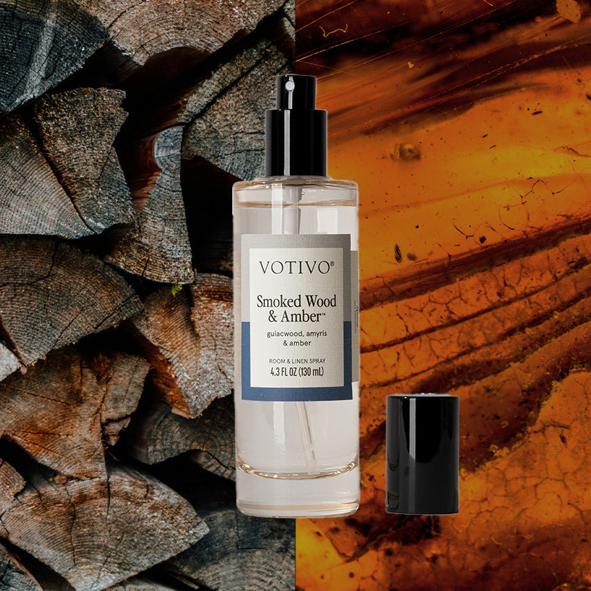 Votivo Smoked Wood & Amber Room & Linen Spray with its cap off, displayed against a background of stacked wood and amber.