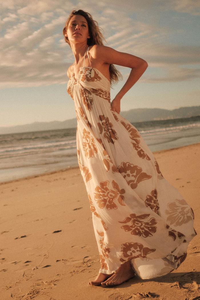 Woman models Promesa Scenic Beauty Halter Maxi Dress, cream & sand floral print, walking on a sunny beach.