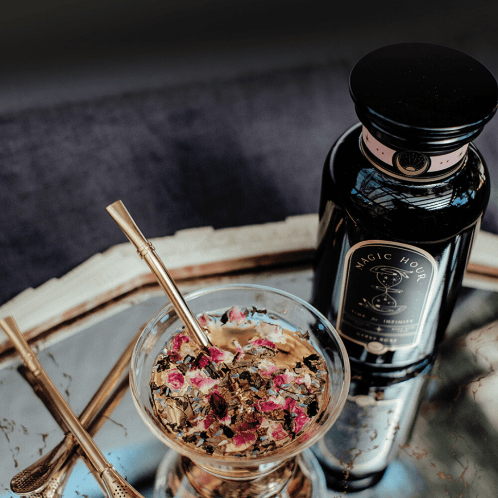 Golden Sips Tea Straw & Strainer Bombilla in rose tea, with a Magic Hour bottle on a reflective tray.