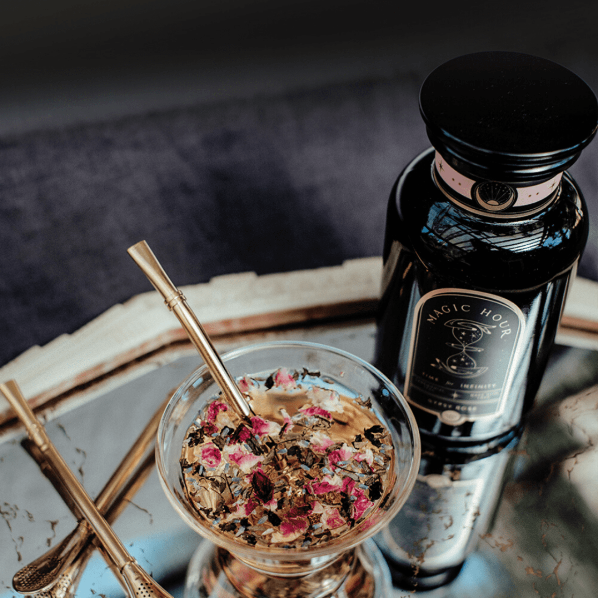Golden Sips Tea Straw & Strainer Bombilla in rose tea, with a Magic Hour bottle on a reflective tray.