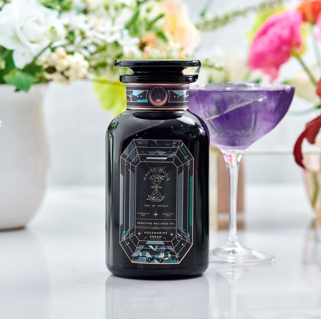 Magic Hour Aquamarine Dream Herbal Adrenal Tonic in a decorative bottle next to a glass of purple tea amidst flowers.