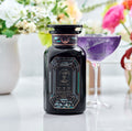 Magic Hour Aquamarine Dream Herbal Adrenal Tonic in a decorative bottle next to a glass of purple tea amidst flowers.