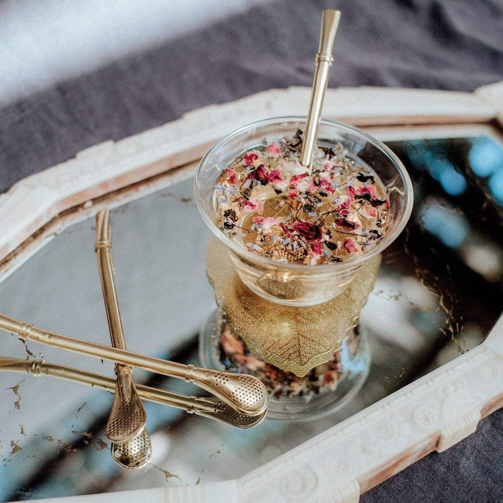 Magic Hour Sips Tea Straw & Strainer Bombilla. Close-up of herbal tea with flower petals and elegant golden bombillas on a mirrored tray.