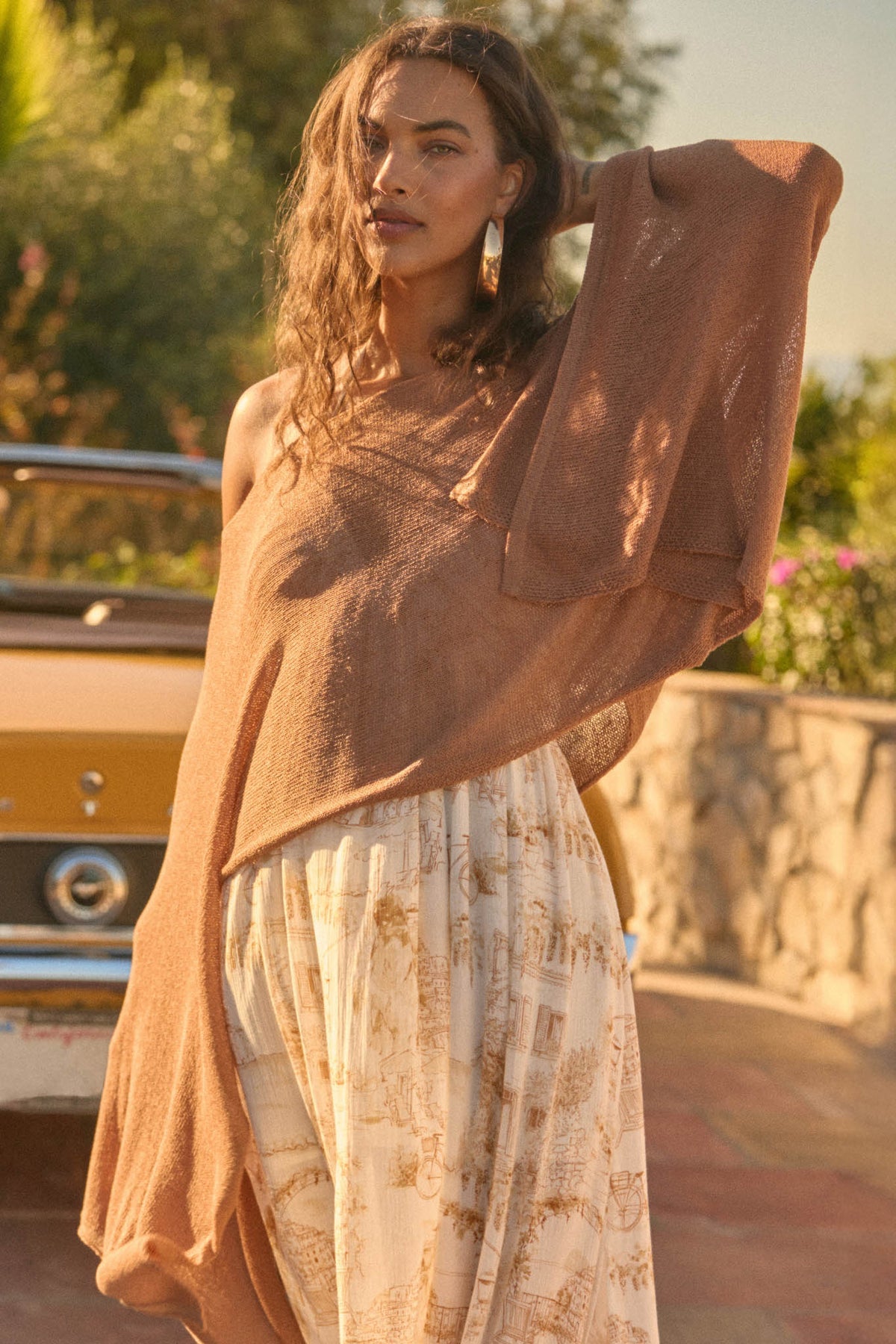 Woman wears Promesa Taupe Sheer to Stay Knit Poncho Sweater & printed maxi skirt outdoors, golden light, vintage car.