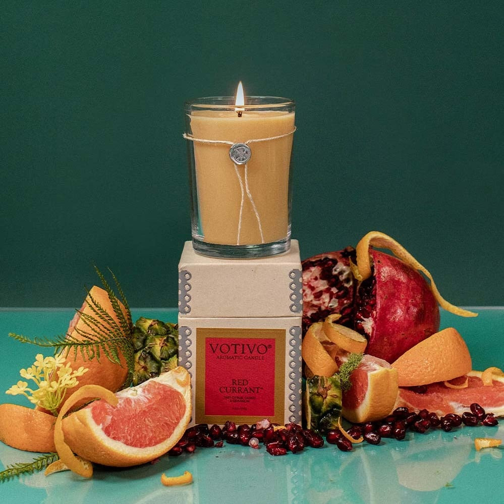 Votivo Red Currant Aromatic Candle, lit in a glass, with its box, surrounded by grapefruit, pomegranate, and orange peels.